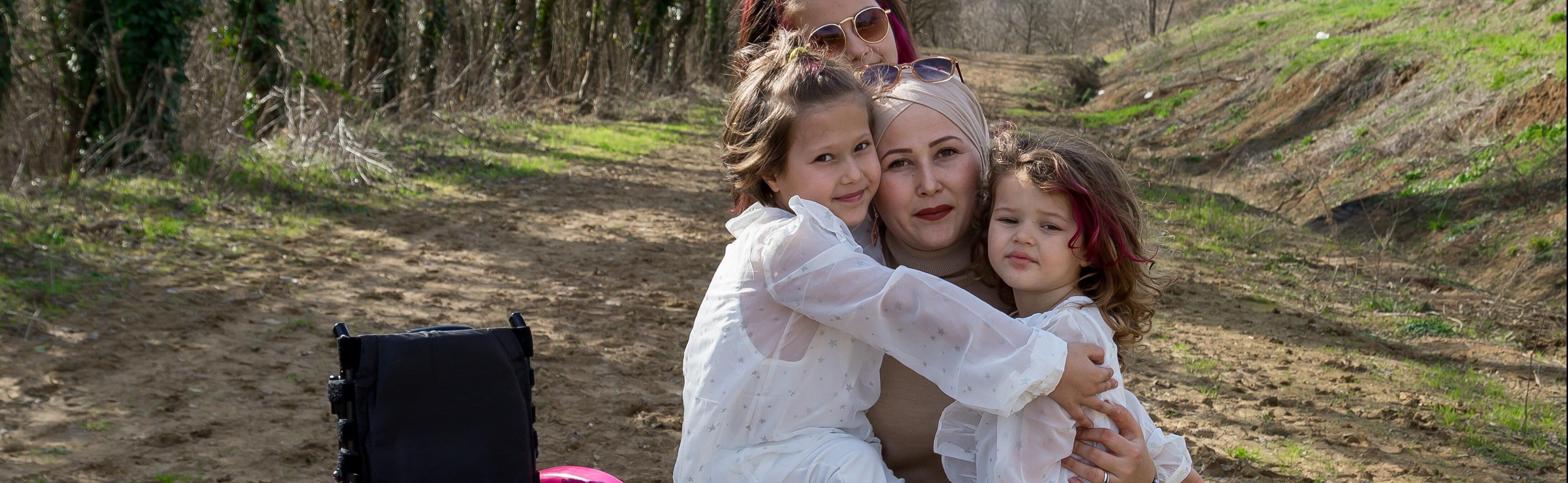 A mother being embraced by her three daughters all wearing white, squatting next to a pink wheelchair outdoors