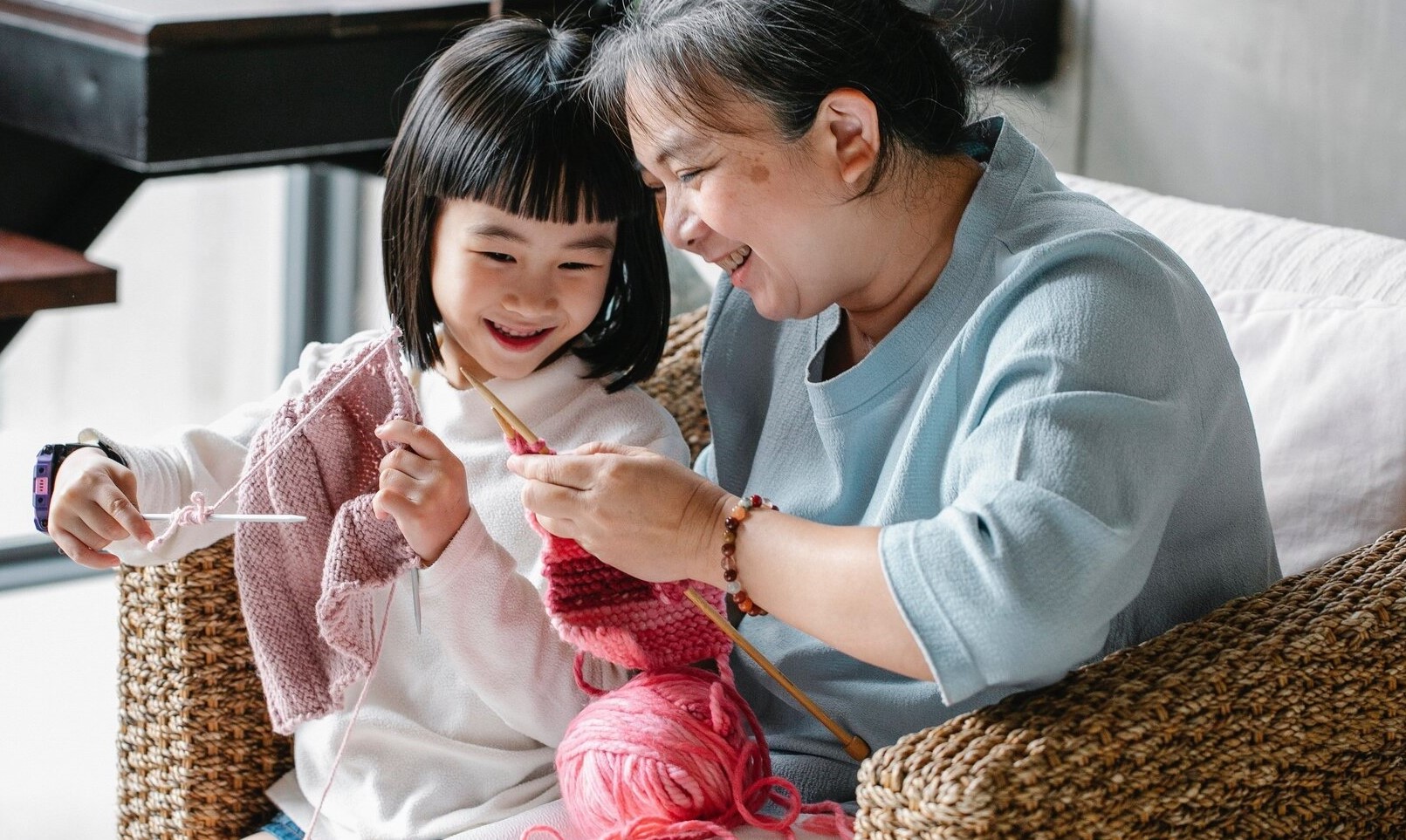A older women smiling with a smaller child seated on her lap in a chair, she teaches the child how to knit with pink yarn