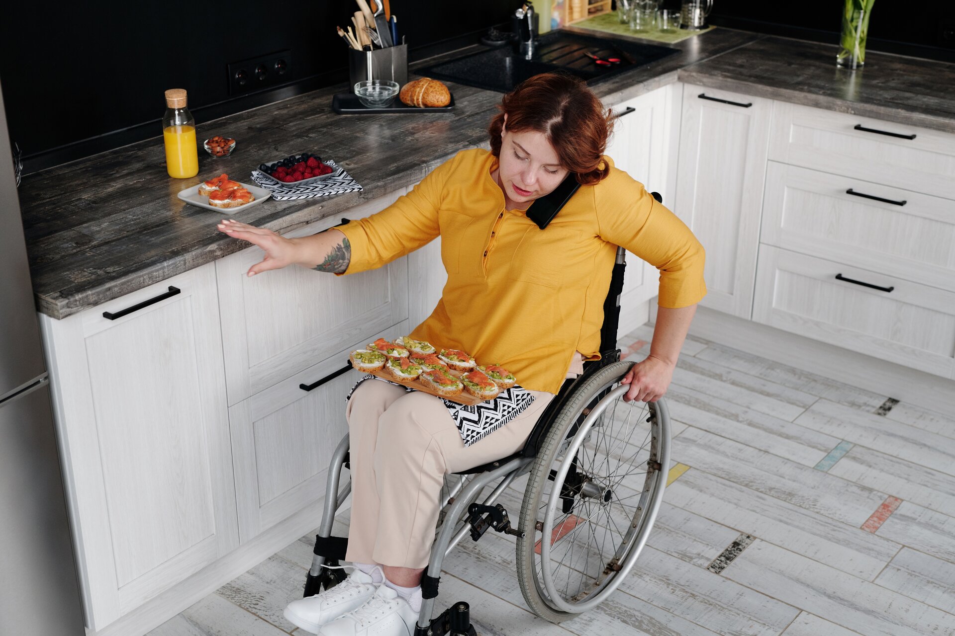 A woman wearing a yellow top and tan pants holding a phone between her ear and her shoulder while she positions food in her lap with one hand while she propels the wheelchair with the other