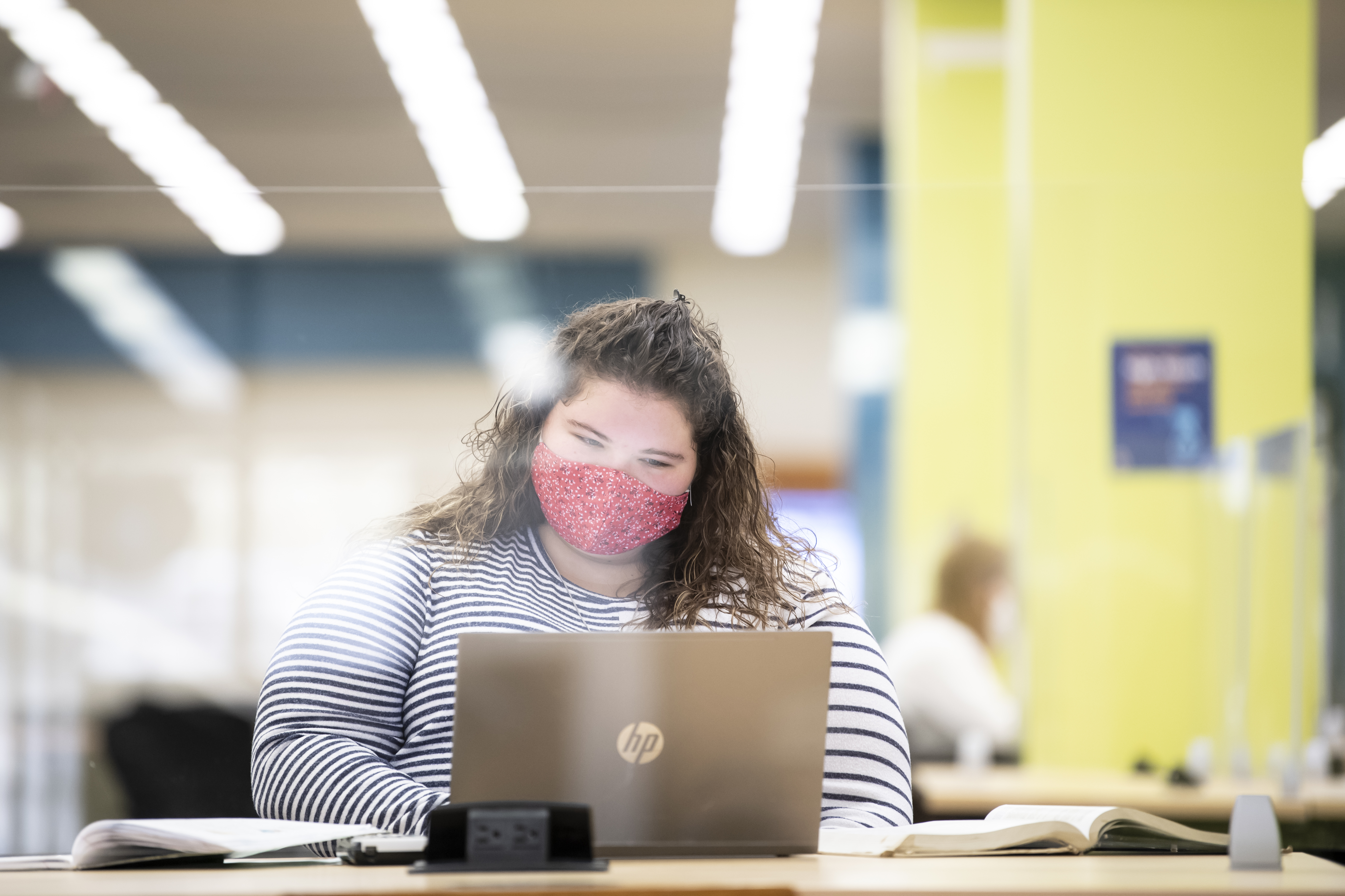 A woman wearing a fabric mask with brown curly hair is typing on her laptop.