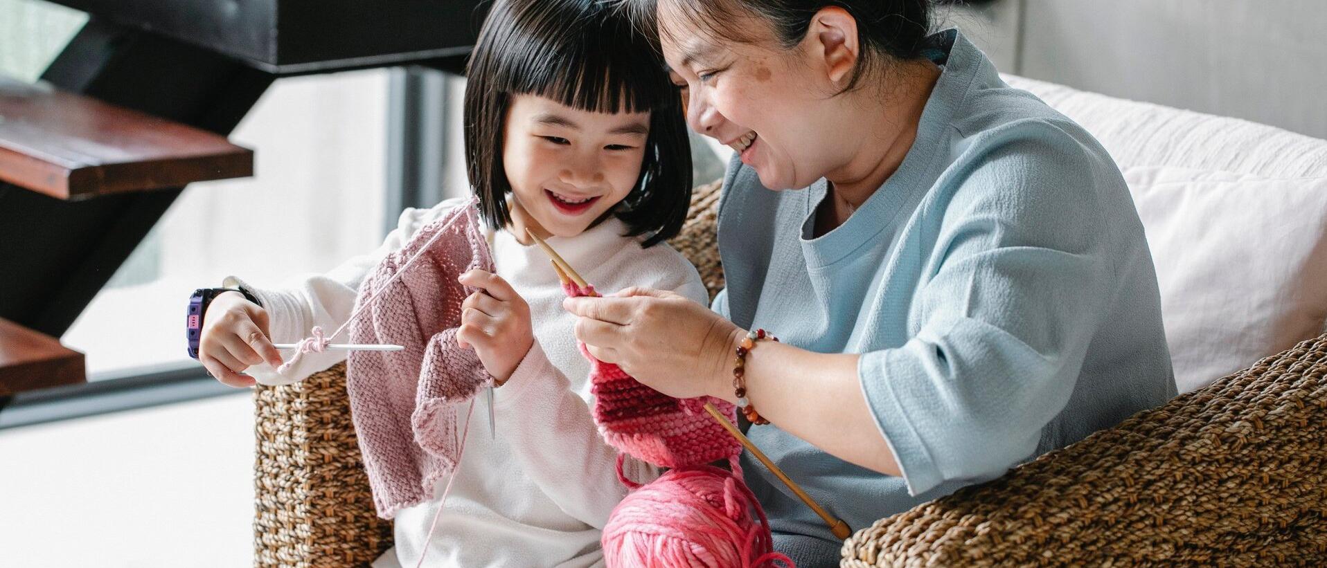 A older women smiling with a smaller child seated on her lap in a chair, she teaches the child how to knit with pink yarn