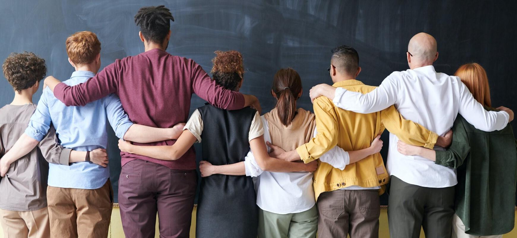Eight diverse people with their backs to the camera holding each other with their arms around their peers backs