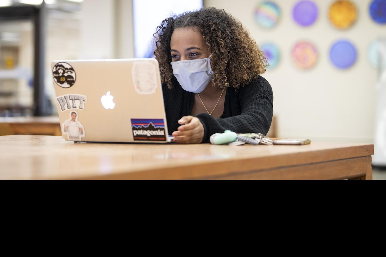 A Black woman with curly hair and a mask on sitting in front of an open laptop.