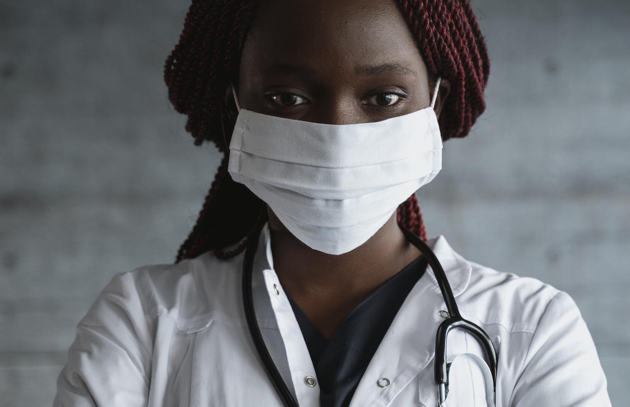 Photo of a woman in white coat and white mask; stethoscope hanging on the back of her neck.