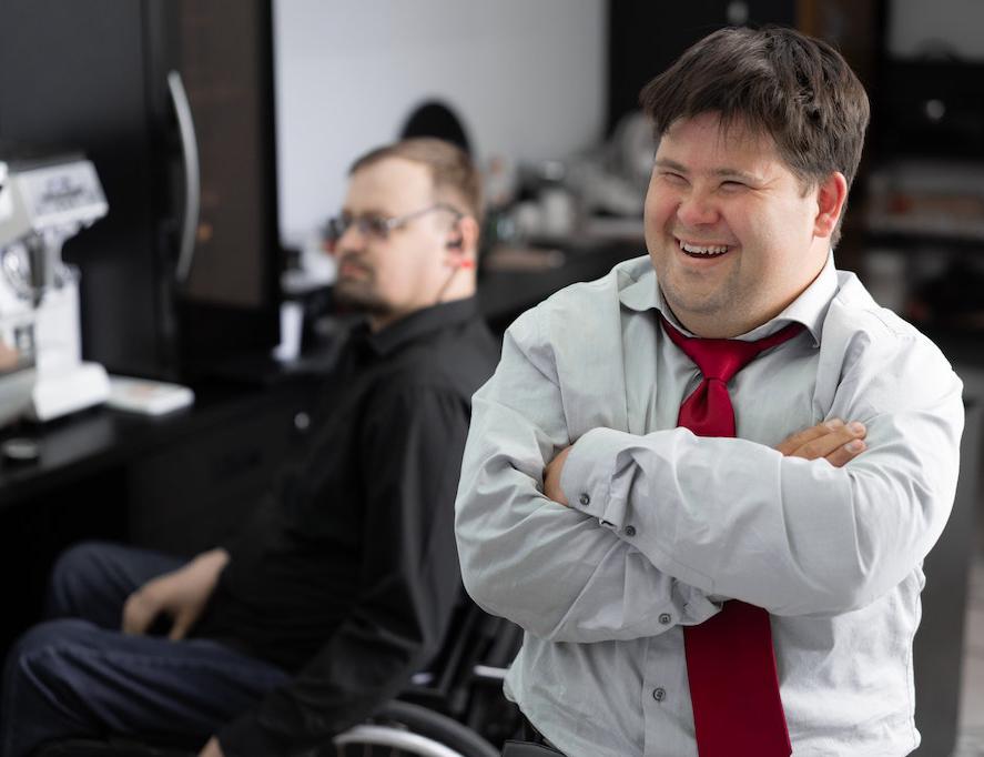 Man with white dress shirt and red tie smiling, another man in the background sits on a wheelchair