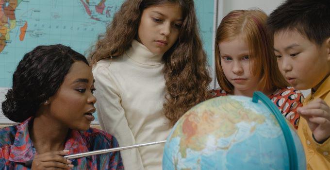 A female teacher holding a pointer directed towards a mobile globe structure surrounded by three children looking at it.