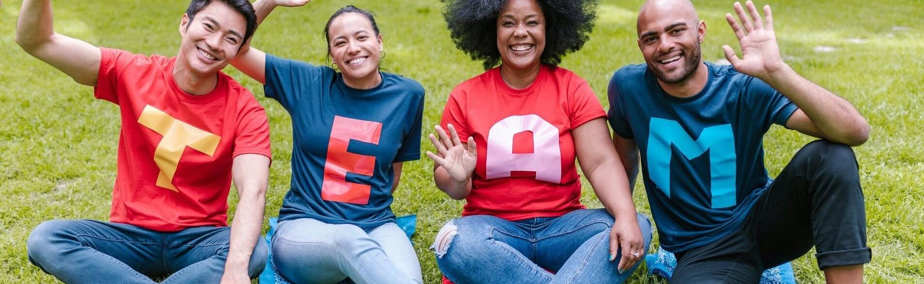 Two Men and two women sitting on the grass outdoors smiling and wearing T-shirts that spell out team