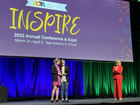 The stage of the AOTF conference. Three white women are on stage, with two in the center with the award and one off to the side.
